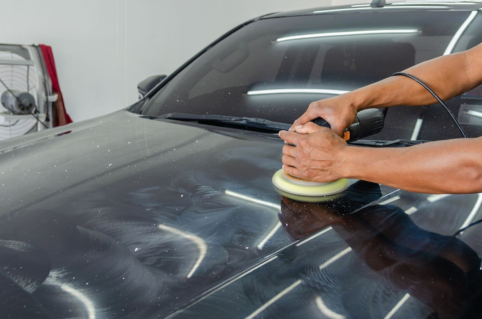 A detailer machine-polishing a black hood under workshop lighting